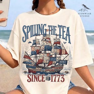 May include: Cream-colored t-shirt featuring a detailed illustration of a sailing ship with red, white, and blue sails. The text "Spilling the Tea" arches above the ship, with "Since 1773" below. The design includes architectural elements and a coastal scene.