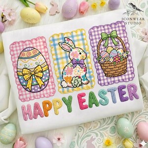 以下が含まれることがあります： イースターをテーマにした白いシャツ。刺繍のデザインが施されています。イースターエッグ、ウサギ、卵のバスケットが、それぞれスカラップフレームの中に描かれています。「HAPPY EASTER」の文字がカラフルな文字で刺繍されています。