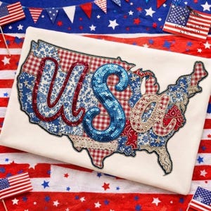 Peut inclure: Un t-shirt crème avec un motif patchwork de la carte des États-Unis. Les lettres "US of A" sont brodées avec des accents rouges, bleus et à paillettes. Le motif incorpore des motifs floraux, vichy et cachemire en rouge, blanc et bleu.