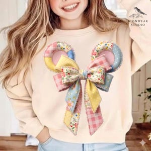 May include: A colorful patchwork bow with circular ear-like shapes. The bow features a mix of floral, gingham, and polka dot patterns in pastel colors like pink, yellow, and blue. The bow is attached to a cream-colored sweatshirt.