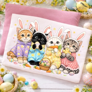 May include: A light pink pillow with a white t-shirt featuring four kittens dressed as Easter bunnies. The kittens wear pastel-colored pajamas and bunny ears, holding Easter eggs, a carrot, and a chick. A basket of eggs and carrots are also present.
