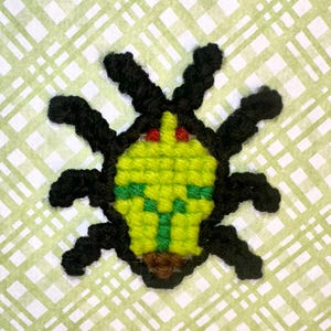 Puede incluir: Un insecto hecho a mano, bordado a punto de cruz, con cuerpo amarillo, detalles verdes y patas y antenas negras. El insecto tiene ojos rojos y boca marrón. El fondo es un patrón de cuadros verde claro y blanco.