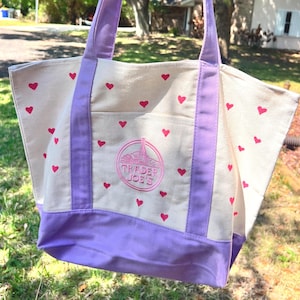 May include: Cream-colored tote bag with a repeating pattern of small pink hearts. The bag features purple accents on the handles, bottom, and a front pocket. The Trader Joe's logo is embroidered on the front pocket.