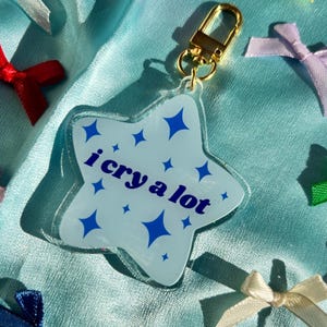 May include: A light blue star-shaped keychain with the text "i cry a lot" in dark blue. The keychain has a gold-colored clasp. The background is a light blue fabric with colorful bows.