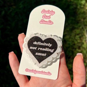 May include: A heart-shaped acrylic pin with a black glitter finish and a white scalloped border. The text "definitely not reading smut" is printed in white. The pin is attached to a white display card featuring "Swirly Owl Studio" text.