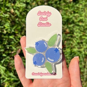 Könnte beinhalten: Eine Acryl-Anstecknadel mit drei glitzernden blauen Heidelbeeren, kleinen dunkelblauen sternförmigen Akzenten und hellgrünen Blättern. Die Anstecknadel ist auf einer weißen, gewölbten Präsentationskarte mit dem Text Swirly Owl Studio befestigt.