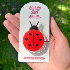 May include: A red ladybug-shaped accessory with black heart details and antennae, held on a white card. The card features the text "Swirly Owl Studio" and the Instagram handle "@swirlyowlstudio".