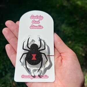 May include: A black widow spider-shaped phone grip with a glittery body and a red hourglass marking. The grip is attached to a white card with the text "Swirly Owl Studio" and the Instagram handle "@swirlyowlstudio".