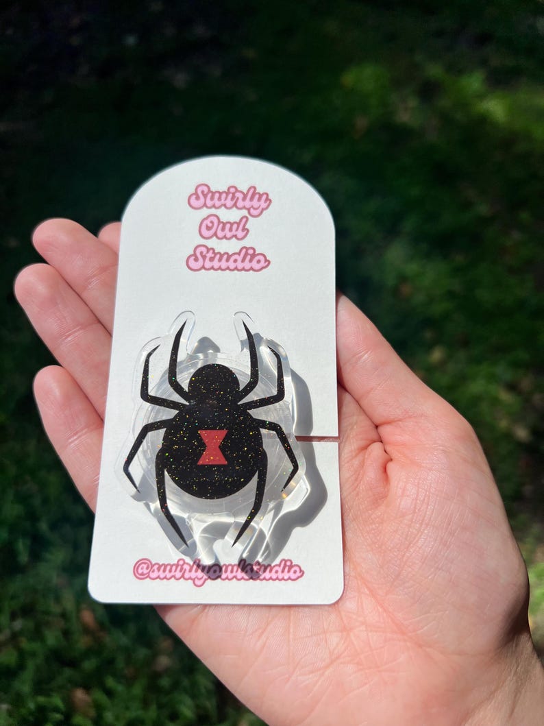 May include: A black widow spider-shaped phone grip with a glittery body and a red hourglass marking. The grip is attached to a white card with the text "Swirly Owl Studio" and the Instagram handle "@swirlyowlstudio".