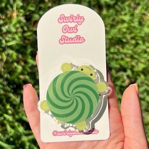 May include: An acrylic pin featuring a green turtle with a spiral shell pattern on a white display card. The card displays the text Swirly Owl Studio at the top and the handle @swirlyowlstudio at the bottom in a pink, rounded font.