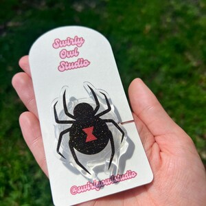 May include: A clear acrylic spider-shaped pin with a black glitter body and a red hourglass marking. The pin is attached to a white card with the text "Swirly Owl Studio" and the Instagram handle @swirlyowlstudio in pink.