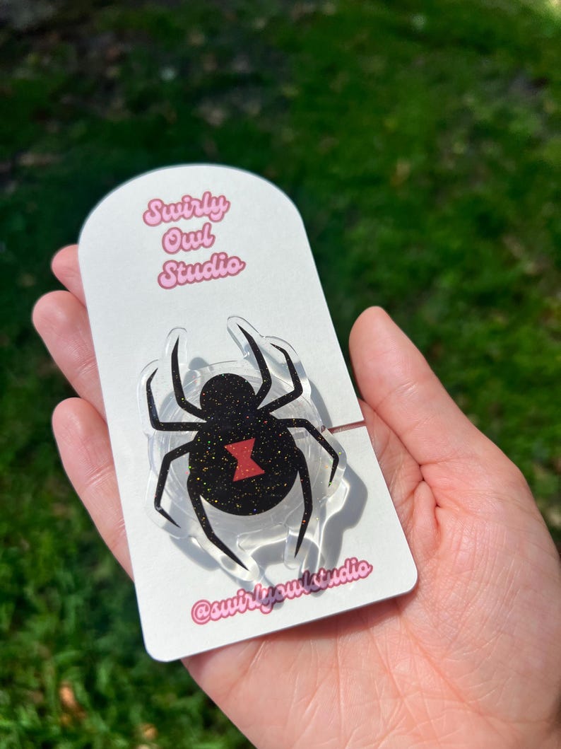 May include: A clear acrylic spider-shaped pin with a black glitter body and a red hourglass marking. The pin is attached to a white card with the text "Swirly Owl Studio" and the Instagram handle @swirlyowlstudio in pink.