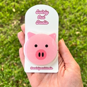 May include: A pink pig-shaped phone grip attached to a white rectangular display card. The card features the text Swirly Owl Studio at the top and @swirlyowlstudio at the bottom in pink script. The pig has black eyes and a pink snout.