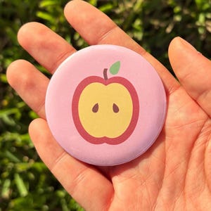 May include: A circular pinback button featuring a light pink background with a centered graphic of a sliced apple. The apple illustration displays a yellow interior, a dark red outer ring, and a small green leaf at the top stem area.