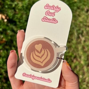 May include: A circular acrylic phone grip featuring a brown coffee cup design with latte art in the shape of a heart and rosetta. The design includes subtle gold glitter accents. It is mounted on a white display card labelled Swirly Owl Studio.