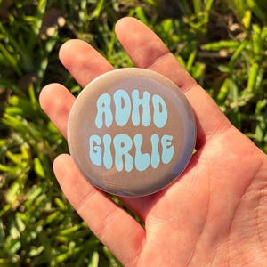 May include: A circular pin-back button with a metallic bronze finish and light blue text that reads ADHD GIRLIE in a wavy, retro-inspired font. The button has a smooth, reflective surface and is circular in shape, featuring bold, rounded lettering.