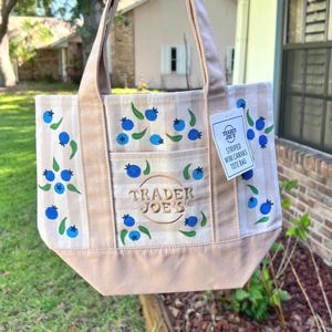 May include: A beige and white striped canvas tote bag featuring a repeating pattern of blue blueberries with green leaves. The bag has a front pocket with the embroidered text TRADER JOE'S and a white tag reading TRADER JOE'S STRIPED MINI CANVAS TOTE BAG.