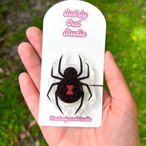 May include: A black widow spider-shaped pin with glitter accents and a red hourglass marking, mounted on a white card. The card features the text "Swirly Owl Studio" and the shop's Instagram handle. The pin is held in a hand.