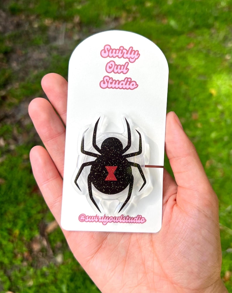 May include: A black widow spider-shaped pin with glitter accents and a red hourglass marking, mounted on a white card. The card features the text "Swirly Owl Studio" and the shop's Instagram handle. The pin is held in a hand.