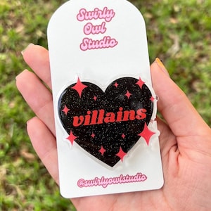 May include: A heart-shaped acrylic pin featuring a black glitter finish with the red text "villains" centered on the front. The design includes several red four-pointed star accents. The pin is attached to a white display card labeled "Swirly Owl Studio".