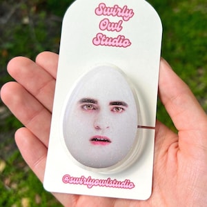 May include: An egg-shaped object with a face printed on it, held on a white card with pink text that reads "Swirly Owl Studio" and "@swirlyowlstudio". The object has a glittery surface.