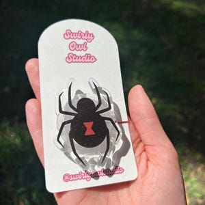 May include: A black widow spider-shaped acrylic pin on a white card. The pin features a black body with a red hourglass marking. The card has the text "Swirly Owl Studio" in pink and the Instagram handle "@swirlyowlstudio".