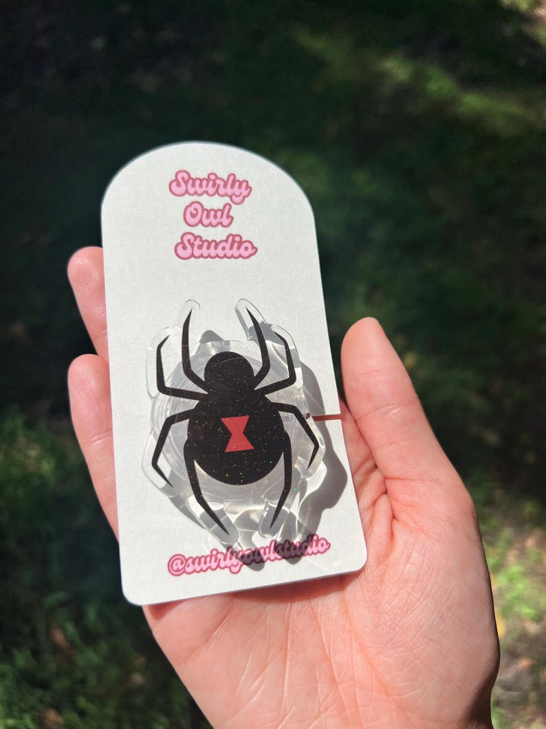 May include: A black widow spider-shaped acrylic pin on a white card. The pin features a black body with a red hourglass marking. The card has the text "Swirly Owl Studio" in pink and the Instagram handle "@swirlyowlstudio".