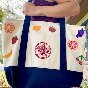 May include: Cream-colored canvas tote bag with navy blue bottom and straps. The bag features colorful illustrations of various fruits, including oranges, strawberries, and grapes. The Trader Joe's logo is printed in red on the front.