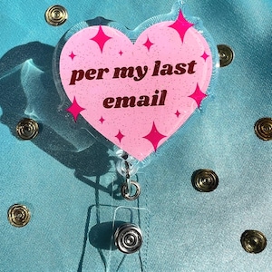 May include: A pink heart-shaped badge reel with the text "per my last email" in brown lettering. The heart has pink starburst accents and a clear plastic reel with a metal clip. The background is a teal fabric.