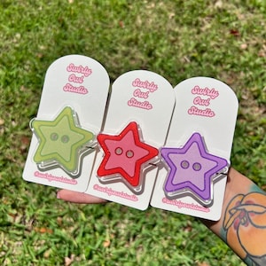 May include: Three star-shaped acrylic pins in light green, red, and purple, each featuring two central button holes. The pins are mounted on white display cards printed with the text Swirly Owl Studio and the handle @swirlyowlstudio.