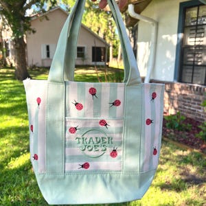 May include: A light green and white striped canvas tote bag featuring a repeating pattern of small red ladybugs. The front of the bag includes an exterior pocket with the embroidered text Trader Joe's inside a circular border in green thread.