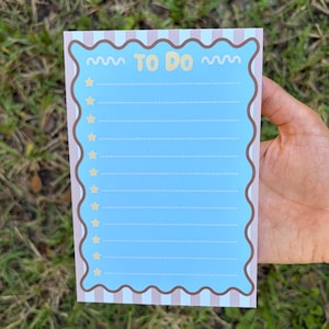 May include: A rectangular notepad with a light blue center featuring the text TO DO in yellow. The notepad has a scalloped brown border and a background of vertical light pink and white stripes. Ten yellow stars mark each dotted line.