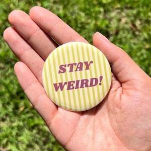 May include: A round pinback button featuring a pale yellow and white vertical striped background with the text STAY WEIRD! printed in a bold, dark maroon serif font across the center of the circular surface.
