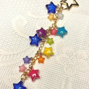 Puede incluir: Un llavero dorado en forma de estrella con una cadena colgante de abalorios de estrella de colores. Los abalorios son en tonos de azul, rosa, amarillo y morado. El fondo es una superficie blanca texturizada.