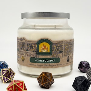 May include: A glass jar candle with a silver lid, labeled "NORSE FOUNDRY." The label features a blacksmith shop illustration with a green and brown color scheme. The candle is surrounded by various polyhedral dice.