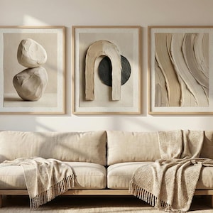 May include: A living room scene featuring a beige sofa with fringed throws, three framed abstract art pieces, and a vase of dried pampas grass. The artwork includes stone-like shapes and textured designs in neutral tones. A wooden side table and window are also visible.