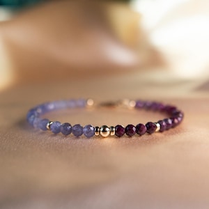 May include: A delicate beaded bracelet featuring a gradient of colors. The bracelet transitions from light blue to deep purple beads, with gold-colored accents. The bracelet is a jewelry item.