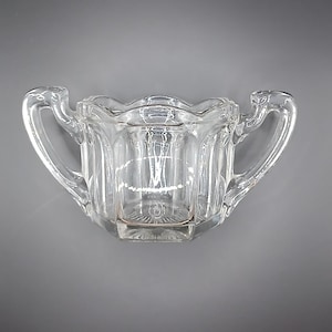 以下が含まれることがあります： 2 つの湾曲したハンドルが付いた透明なガラスの砂糖入れ。ボウルはスカラップの縁と垂直のリブのディテールがあります。ボウルの底には装飾的な円形の模様があります。砂糖入れは空で、灰色の背景にあります。