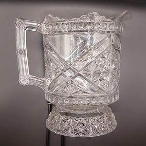 May include: A clear, cut-glass jug with a handle and a decorative base. The jug features a diamond and starburst pattern, with a scalloped rim. The handle is rectangular and has decorative detailing. The jug is likely used for serving beverages.