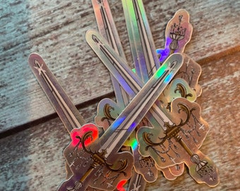May include: A stack of iridescent holographic stickers featuring a sword with a chandelier-style handle. The stickers are on a wooden surface.
