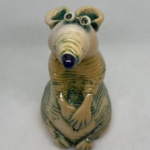 以下が含まれることがあります： 風変わりなデザインのセラミックマウスの置物。マウスは長く縞模様の鼻、大きな耳、丸い体を持っています。配色はベージュ、グリーン、ブルーで、光沢仕上げです。マウスは足を組んで座っています。