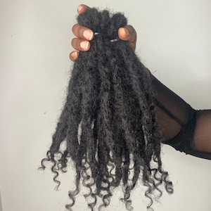 May include: A hand holding a bundle of dark, curly hair extensions. The hair is a deep black colour with tight, defined curls at the ends. The extensions appear to be made of natural hair.