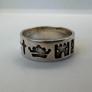 Puede incluir: Un anillo de plata con símbolos grabados, incluyendo una cruz, una corona y un castillo. El anillo tiene una banda ancha y una apariencia ligeramente desgastada. Los símbolos son oscuros sobre la plata. El diseño sugiere un tema histórico.