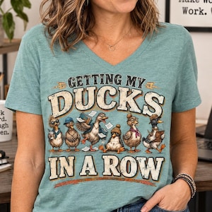 May include: A heathered teal V-neck t-shirt featuring a graphic of six cartoon ducks with various office accessories. The text reads GETTING MY DUCKS IN A ROW in a distressed, bold font. The design includes ducks wearing glasses, hats, and holding papers.