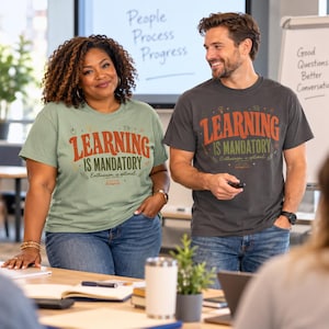 May include: A sage green t-shirt and a charcoal gray t-shirt, both featuring the text "LEARNING IS MANDATORY" in large orange letters, with "Enthusiasm is optional" in smaller script below, accompanied by small icons of books, pencils, and stars.