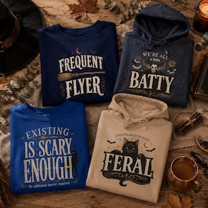 May include: A collection of four garments featuring Halloween-themed graphics: a navy sweatshirt with 'FREQUENT FLYER' and a broom, a dark blue hoodie with 'WE'RE ALL A LITTLE BATTY' and a skull, a blue sweatshirt with 'EXISTING IS SCARY ENOUGH', and a beige hoodie with 'UNAPOLOGETICALLY FERAL' and a black cat.