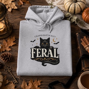 May include: A light gray hooded sweatshirt featuring a graphic print of a black cat with yellow eyes, surrounded by small stars, a crescent moon, and two bats. The text "UNAPOLOGETICALLY FERAL" is printed in a decorative font above and below the cat.