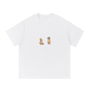 May include: White t-shirt featuring a minimalist illustration of two cartoon teddy bears. One bear wears a crown, the other a cap. The design is centered on the shirt, offering a clean and simple aesthetic.