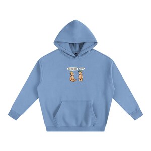 May include: A light blue hoodie with a kangaroo pocket and a drawstring hood. The front features a cartoon illustration of two teddy bears with speech bubbles. One bear wears a hat and the other a cap. The text in the speech bubbles is not clear.