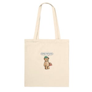 May include: Cream-colored tote bag with a cartoon illustration of a teddy bear wearing a green cap and holding a pink purse. The bear has a speech bubble that says "luckily I have purse."